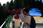 Hiker at a vantage point of the Rax, looking through a telescope. In the background there is a slope with tall conifers.