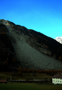 A mountain with a scree slope, below that a road, a hut and to the right a house. Larches in autumn dress. Above blue sky.