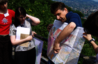 The focus is on some people. A man holds a geological map in hand, pointing at them and said so. In the background is a wooded slope and a remote village.