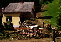 A house which is missing the right half, the roof is intact. In the foreground debris, rubble and devastation. Right in the background a grassy slope and on its left side a breakaway edge and the course of the landslide.