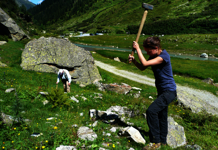A stream is flowing through the center of a valley bordered by green meadows. Rock boulders of various sizes lie on the valley floor. A standing rman in the background is looking at the ground, a woman in the foreground is swinging a big hammer to cut off