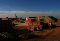 A large square with attached pour requested mound in the background goes a yellow loader. Everywhere there are several pallets. Bricks are stacked on it. Some have a red and white packaging.