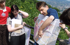 The focus is on some people. A man holds a geological map in hand, pointing at them and said so. In the background is a wooded slope and a remote village.