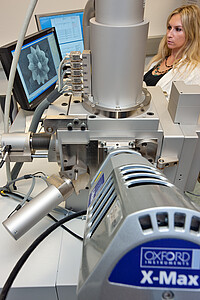 Ein Rasterelektronenmikroskop, links dahinter ein Bildschirm, rechts dahinter eine Dame.