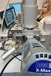 Ein Rasterelektronenmikroskop, links dahinter ein Bildschirm, rechts dahinter eine Dame.