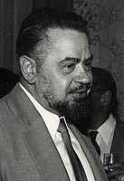 A man with a beard, tie and jacket. Right behind a man with hidden face and a glass in his hand.
