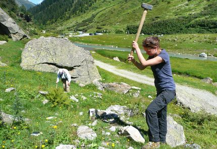 A stream is flowing through the center of a valley bordered by green meadows. Rock boulders of various sizes lie on the valley floor. A standing rman in the background is looking at the ground, a woman in the foreground is swinging a big hammer to cut off