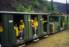 A small electric locomotives with Wagon without windows. Children look out of the open doors. They wear yellow jackets and helmets.