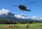 A lawn with bushes and forested mountains in the background. Clear skies with some clouds. Five people are on the lawn. Two are beside a large, almost 6m long meter. Two use a film camera. An army soldier. A helicopter hovers over them with a tow cable.