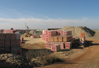 A large square with attached pour requested mound in the background goes a yellow loader. Everywhere there are several pallets. Bricks are stacked on it. Some have a red and white packaging.